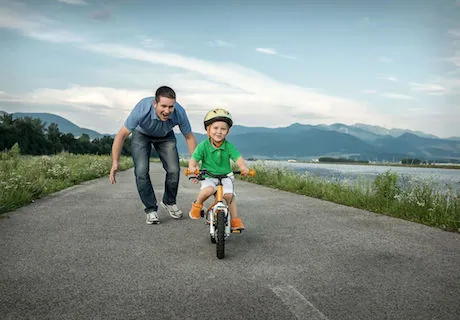 Adult man jogging alongside young child on small bicycle on rural road with mountains and lake in background